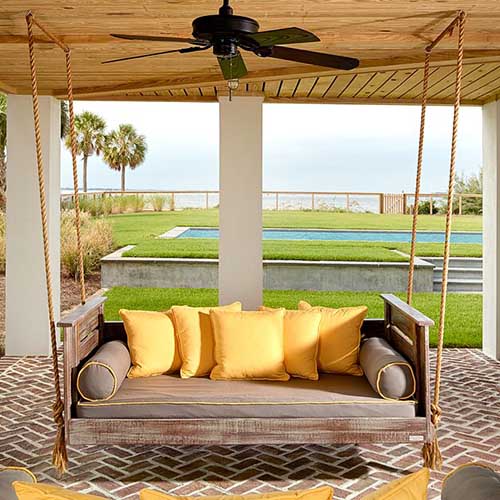 An open sided, covered porch with a wooden bench seat hanging from the ceiling overlooking a garden scene with a pool and the ocean in the background.