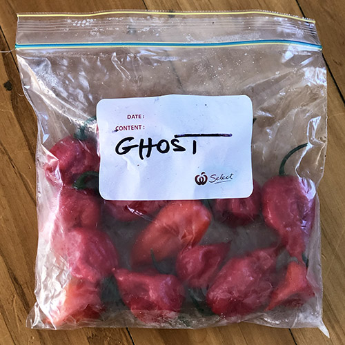 A close up of a small bag of red 'Bhut Jolokia' fruits that are labelled and frozen, set on a wooden surface.