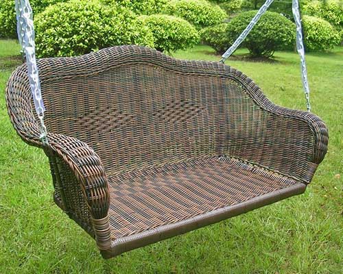 A close up of a brown wicker porch swing hanging from metal chains, set up in the garden with bushes and shrubs in the background.
