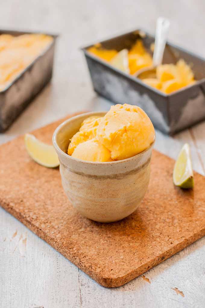 A vertical picture of a small ceramic bowl with a fresh homemade mango, lime, and cream cheese sherbet, set on a cork board on a wooden surface.