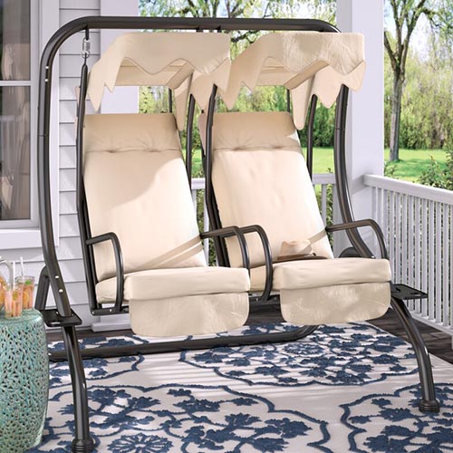 A metal-framed, two seater porch swing with beige cushions and canopy on a deck outside a white wooden home.