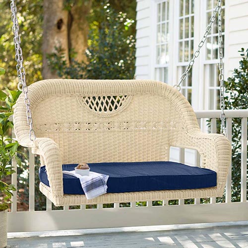 A wicker hanging bench seat with rounded edges and a dark blue cushion on a deck, with the side of a house and garden scene in the background in soft focus.