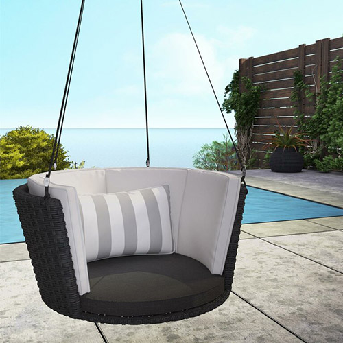 A circular hanging seat, in black wicker with white striped cushions hanging over a concrete patio with a swimming pool and the ocean in the background.