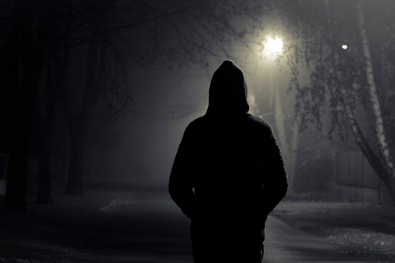 A back view of a sinister man wearing a hoodie walking along a street in the nighttime with a streetlamp for light.