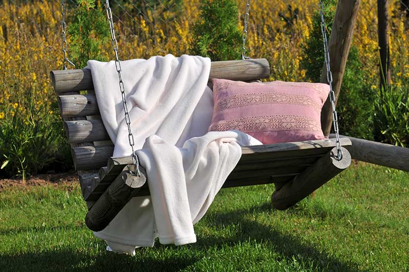 A close up of a rustic wooden hanging bench seat with a pink cushion and beige throw, pictured in the garden in light sunshine.