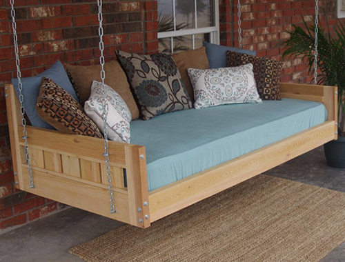 A wooden bench seat with a blue mattress and colorful scatter cushions hangs from chains on a porch with a brick building in the background.