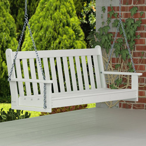A white bench hanging from chains on a white wooden porch with a brick building and conifer trees in soft focus in the background.