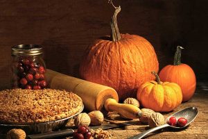 A close up of an autumnal kitchen scene, with a pie, a rolling pin, unshelled walnuts, and three pumpkins on a rustic wooden surface on a dark background.