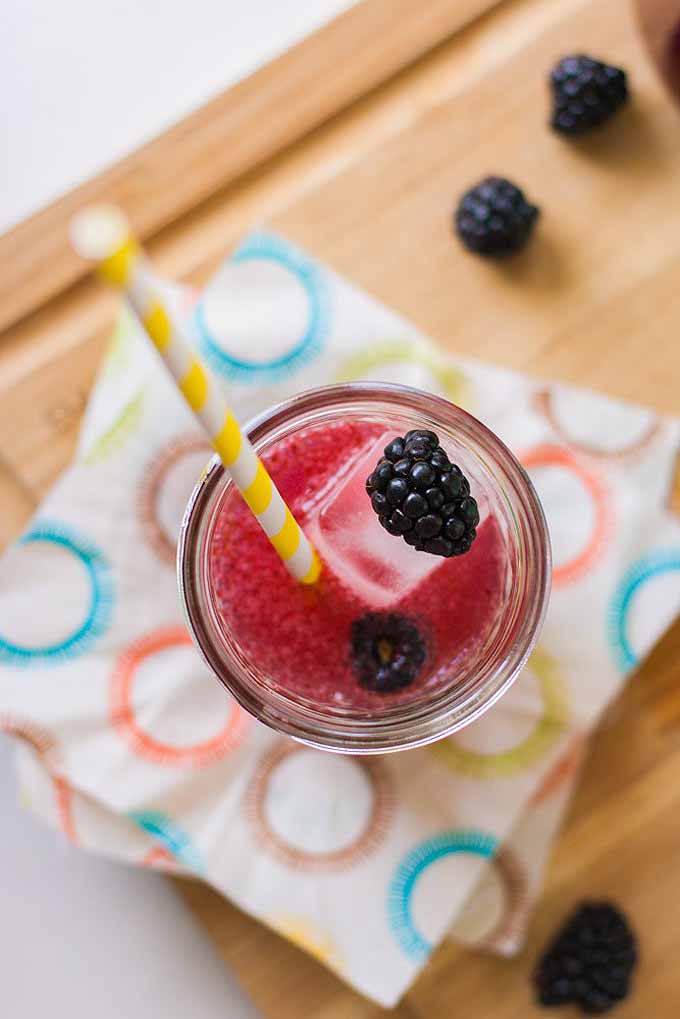 A vertical top down picture of a small glass of a blackberry lemon cocktail set on a colorful paper towel on a wooden surface.