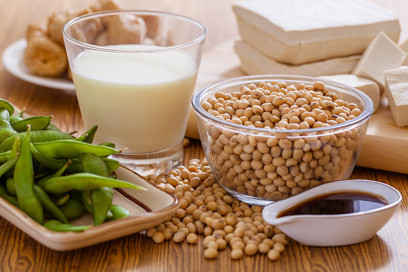 A close up of a variety of different soy-based products, including soy beans, edamame, soy milk, tofu and tempeh, set on a wooden surface.