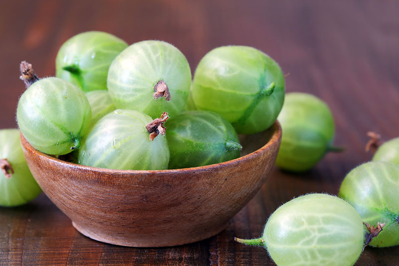 A close up of a small wooden bowl with green gooseberries spilling out onto a wooden surface.