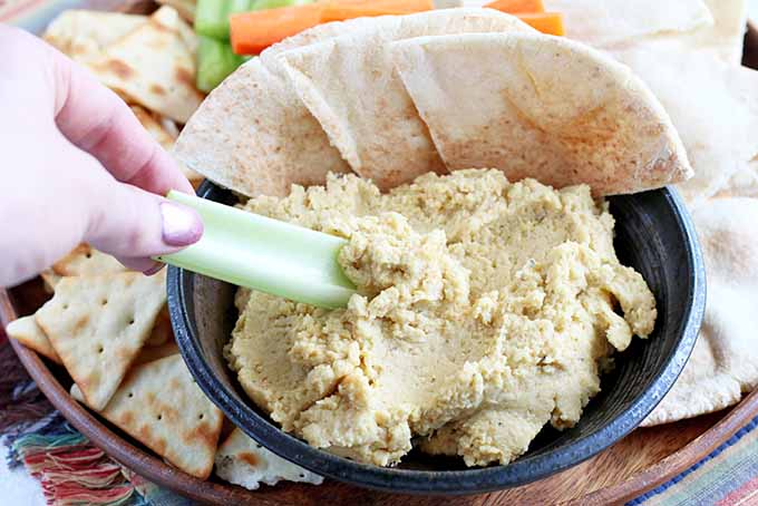 A close up of a bowl of roasted garlic hummus set on a wooden tray with freshly baked flatbreads, and a hand to the left of the frame.