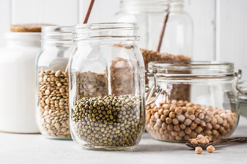 A close up of jars containing various dried lentils and beans, as part of a plant-based diet, set on a white surface, with a white wooden wall in the background.
