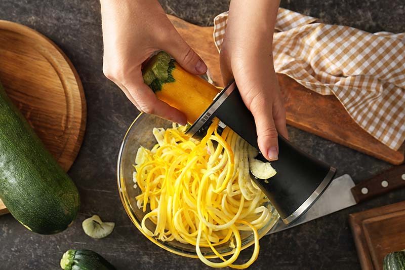 A close up of two hands from the top of the frame, using a spiralizer to make zucchini noodles, in a small metal bowl, over a dark gray surface.