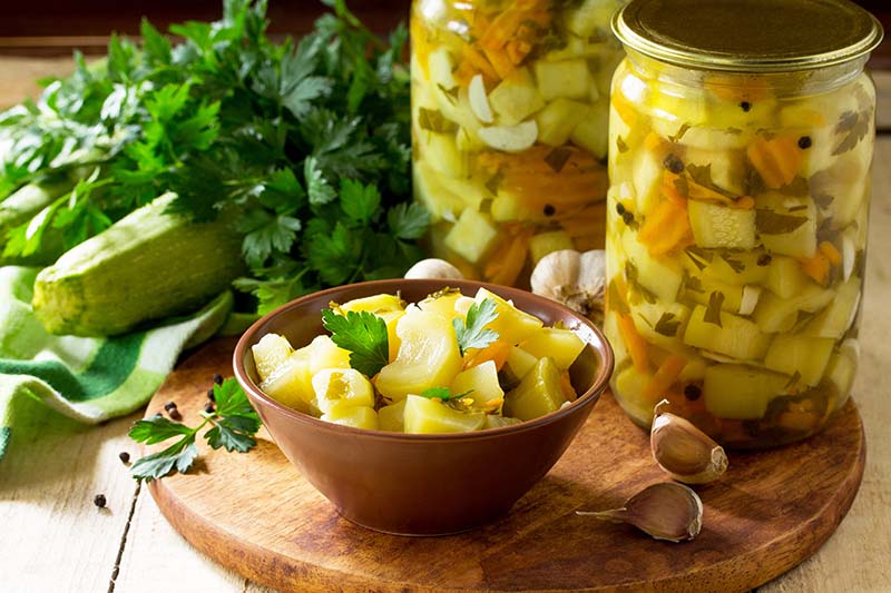 A close up of a small bowl and two glass jars with pickled zucchini set on a wooden surface, with herbs in soft focus in the background.
