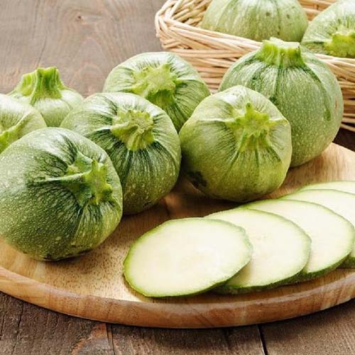 A close up of a wooden chopping board with round zucchini squash, some whole and one sliced into rounds. The variety is light green with light flecks and is called 'Ronde de Nice.' In the background is a wicker basket.