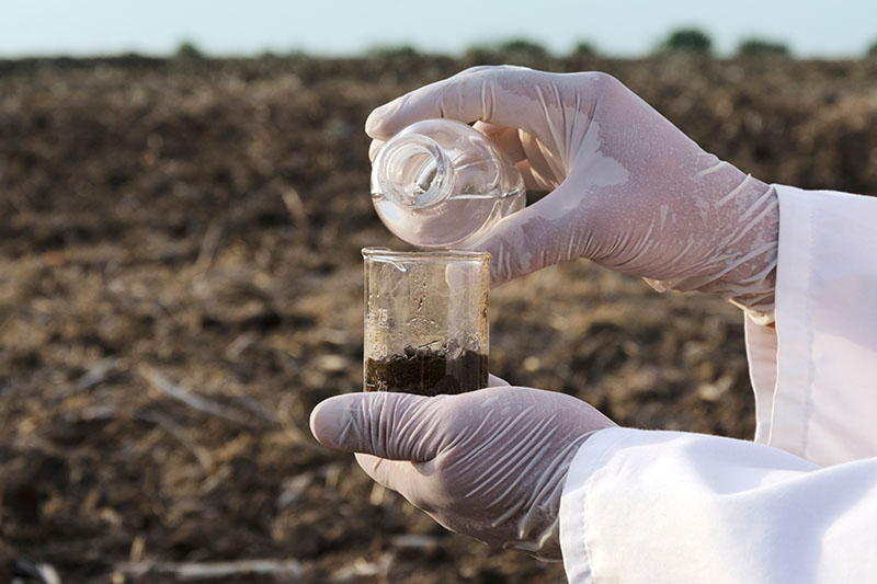 A close up of gloved hands from the right of the frame pouring liquid into a beaker of soil to test the nutrient content.