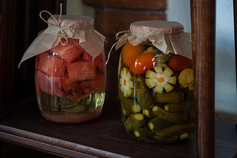 A close up horizontal image of two glass jars containing canned vegetables and fruits set on a wooden shelf.