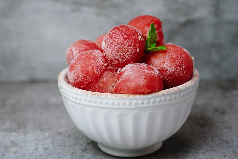 A close up horizontal image of a small white bowl containing round scoops of frozen watermelon set on a gray stone surface.