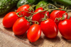 A close up of bright red, ripe 'Roma' tomatoes, still attached to the vine, set on a wooden surface with herbs in soft focus in the background.