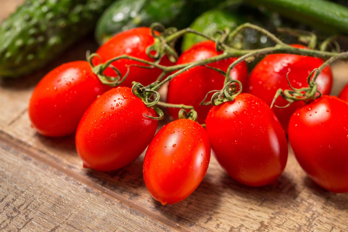 A close up of bright red, ripe Roma tomatoes, still attached to the vine, set on a wooden surface with herbs in soft focus in the background.