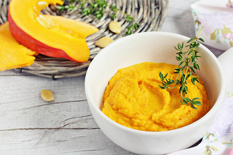 A close up of a small white bowl containing homemade pumpkin puree topped with a sprig of thyme, set on a wooden surface, with slices of squash on a basket in the background.