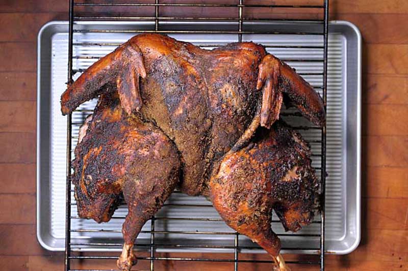 A close up, top down picture of a spatchcock chicken on a grill placed over a metal tray set on a wooden surface.