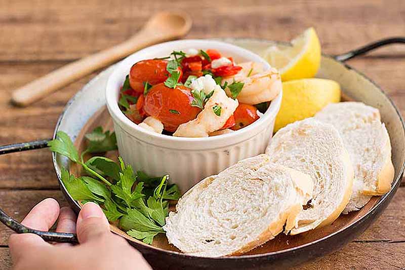 A close up of a ramekin of roasted cherry tomatoes with shrimp and feta served with bread slices, lemon, and coriander on a wooden table background.