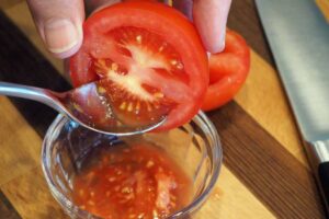 A close up horizontal image of a hand from the top of the frame holding a tomato that has been cut in half, and a spoon scooping out the seeds.