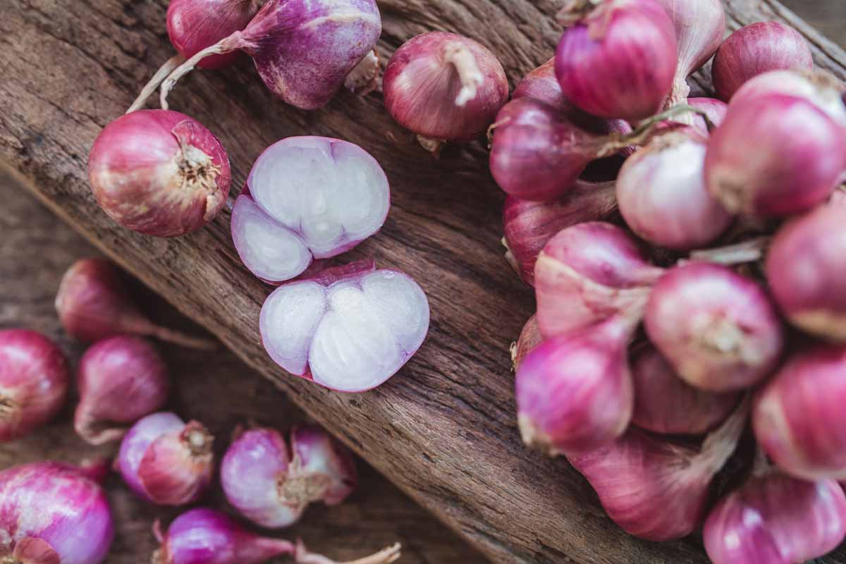 A close up of a pile of red shallots with thin, papery skin set on a wooden surface. To the center of the frame, one of the bulbs has been cut in half, revealing the separate cloves.