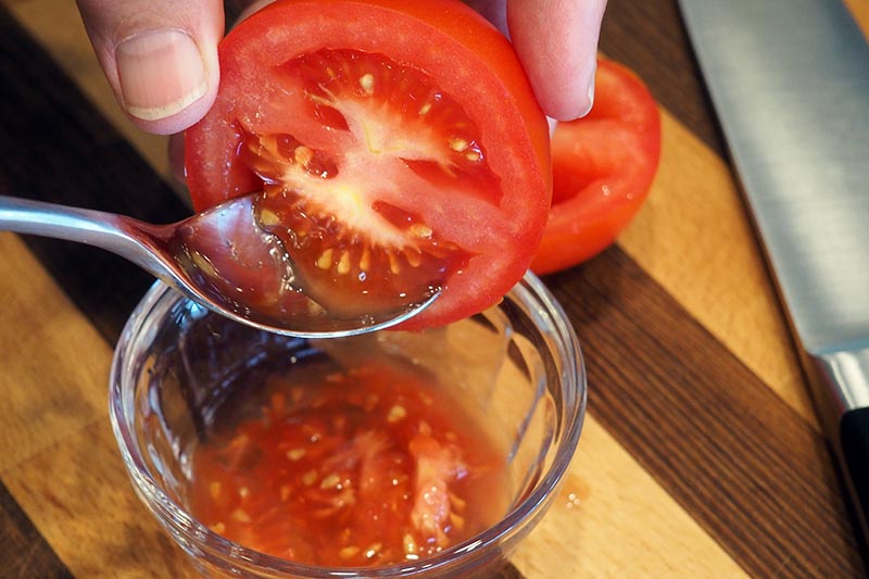 A close up horizontal image of a hand from the top of the frame holding a tomato that has been cut in half, and a spoon scooping out the seeds. In the background is a wooden surface and a metal knife.