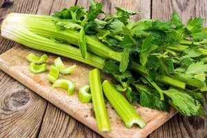 A close up horizontal image of a freshly harvested celery plant set on a wooden chopping board. In the foreground are chopped stalks.