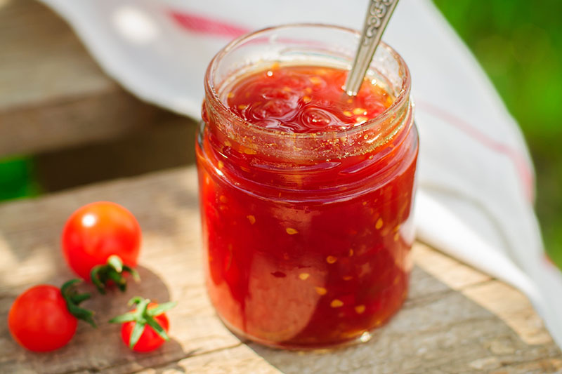 A close up horizontal image of a glass jar with a spoon standing up in it, filled with fresh, homemade tomato jam, set on a wooden surface pictured in bright sunshine on a soft focus background.