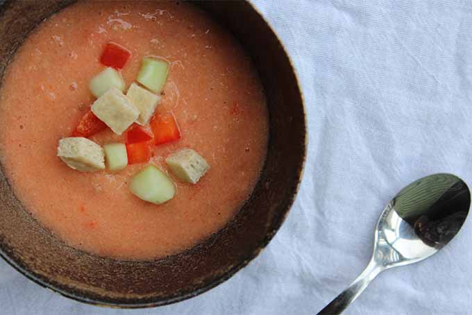 A close up horizontal image of a bowl of gazpacho soup set on a white fabric surface with a silver spoon to the right of the frame.