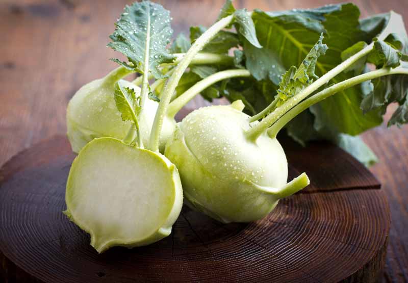 A close up horizontal image of freshly harvested and cleaned kohlrabi with two whole in the background and one sliced in half to the front, set on a wooden chopping board on a wooden counter.