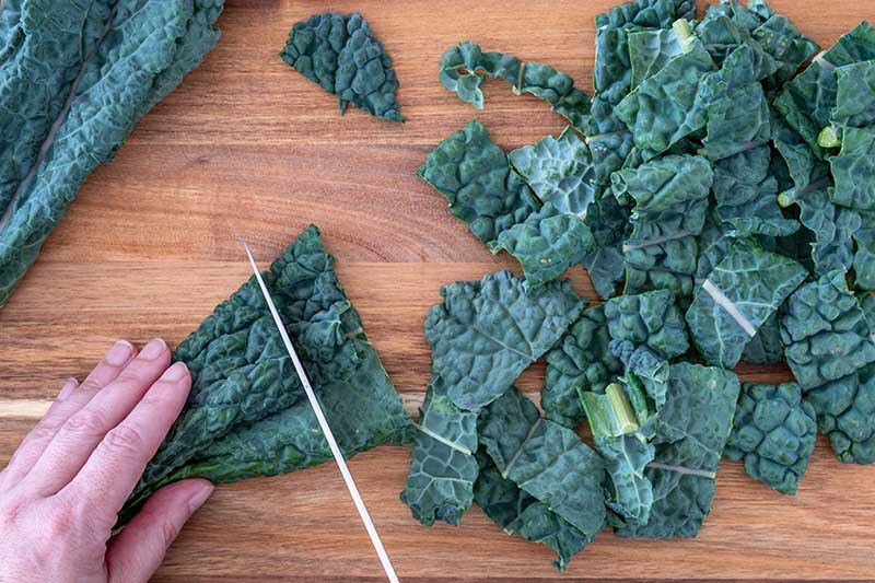 A close up horizontal image of a hand to the left of the frame chopping leafy greens into small chunks on a wooden surface for cooking.