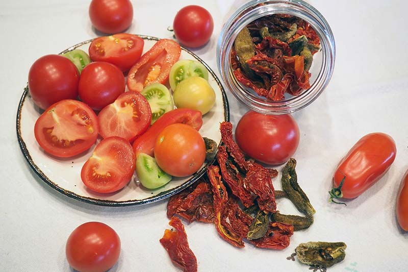 Uma imagem horizontal aproximada de um prato de tomates cultivados em casa recém-colhidos à esquerda do quadro e desidratados em uma jarra à direita, sobre uma superfície branca.
