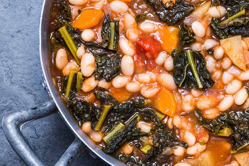 A close up top down horizontal image of a metal saucepan of a beans and greens dish freshly cooked set on a gray surface.