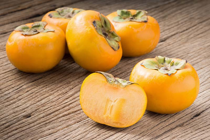 A close up horizontal image of bright orange American persimmons set on a wooden surface, with one cut in half to show the flesh inside.