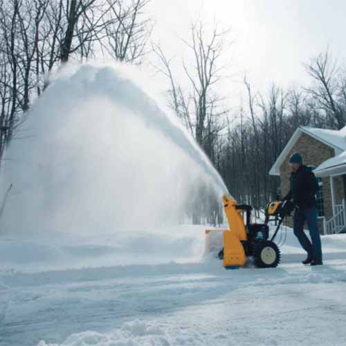 A square image showing the yellow and black Cub Cadet being used to clear snow in the garden with a house and trees in the background.