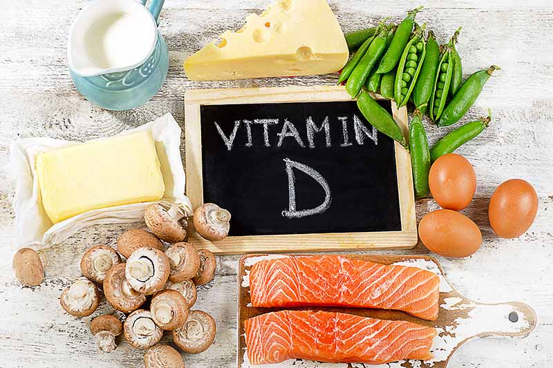 A close up horizontal image of a chalkboard sign saying Vitamin D with a variety of foodstuffs around it.