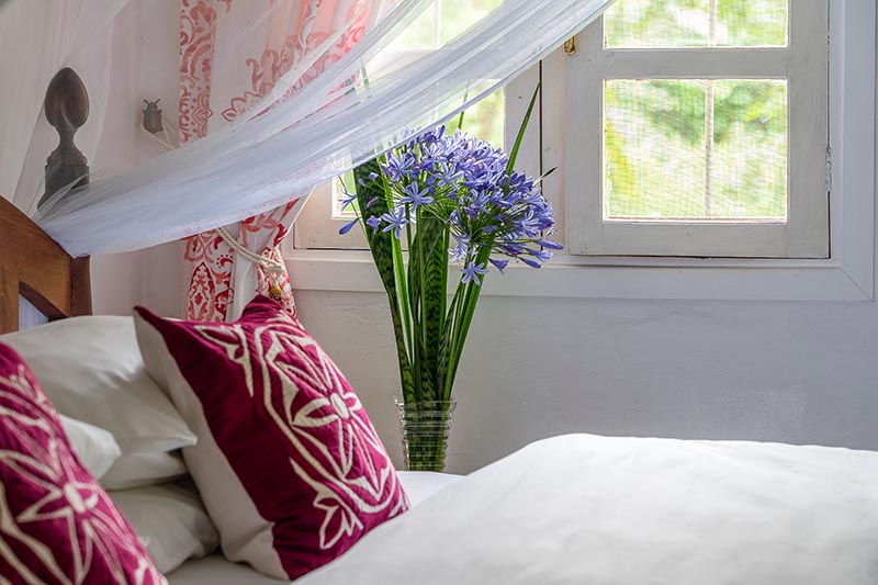 A close up horizontal image of a bedroom with white sheets, dark red cushions, and a vase of bright blue cut flowers on a sidetable. In the background is a window with a garden scene behind.