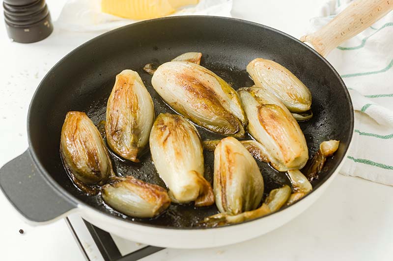 A close up horizontal image of a frying pan filled with braised Belgian endive set on a white surface.
