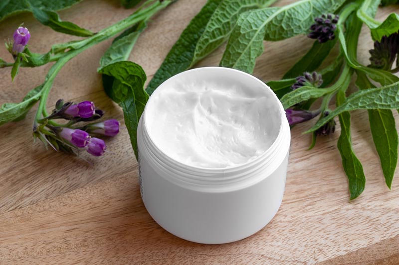 A close up horizontal image of a small white plastic pot with freshly made comfrey salve set on a wooden surface with leaves and flowers in the background.
