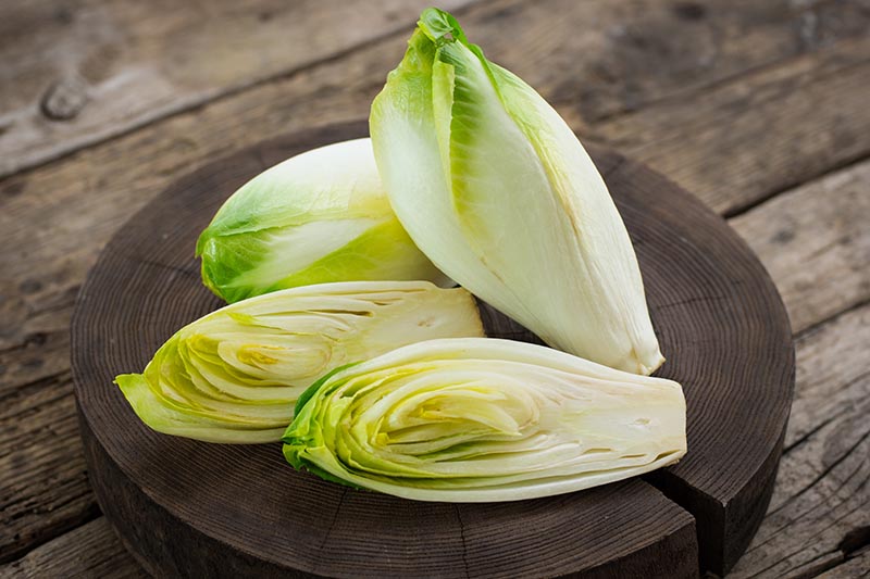 A close up horizontal image of three heads of Belgian endive with one cut in half set on a wooden chopping board on a dark brown surface.