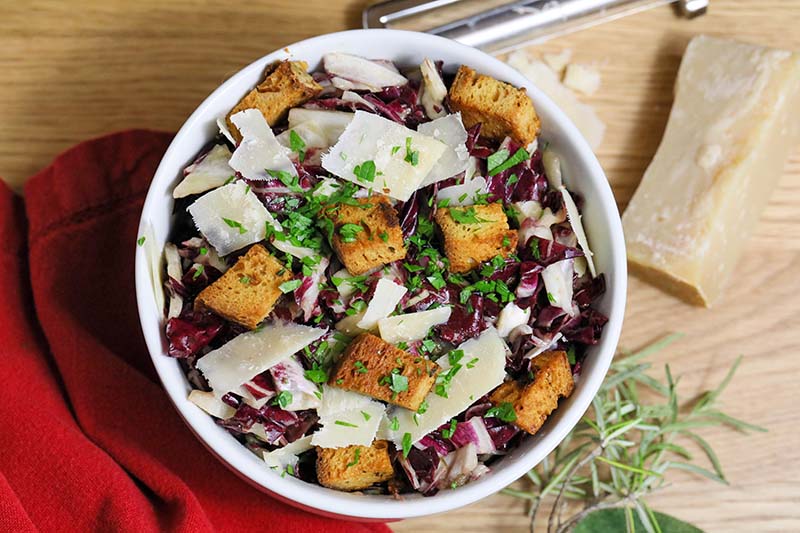 A close up top down image of a white bowl of salad topped with parmesan cheese and croutons set on a wooden surface.