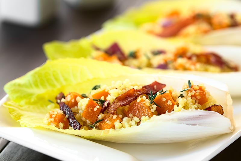 A close up horizontal image of witloof leaves stuffed with couscous, bacon, and vegetables pictured on a soft focus background.