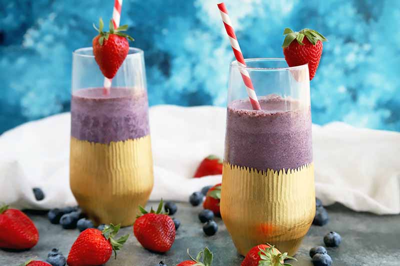 A close up horizontal image of two glasses of freshly made smoothies with fruits scattered around them.