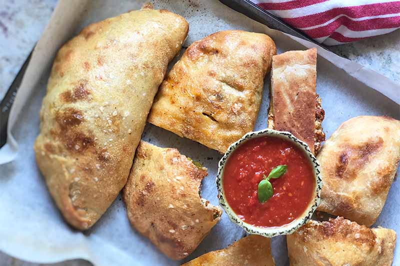 A close up horizontal image of freshly baked calzone pizza slices on a baking sheet with a bowl of fresh tomato sauce in the center.