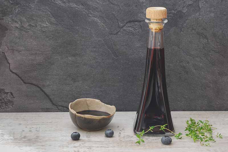 A horizontal image of a glass bottle with a cork stopper filled with homemade blueberry vinegar set on a wooden surface with a sprig of thyme and fresh berries scattered around. To the left of the frame is a small bowl.
